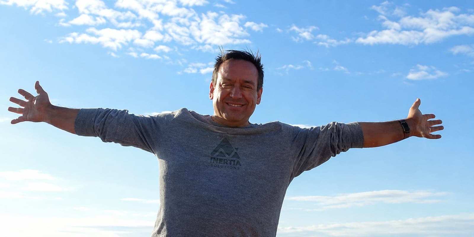 James Moss, arms wide under a blue sky, in an Inertia Solutions shirt.