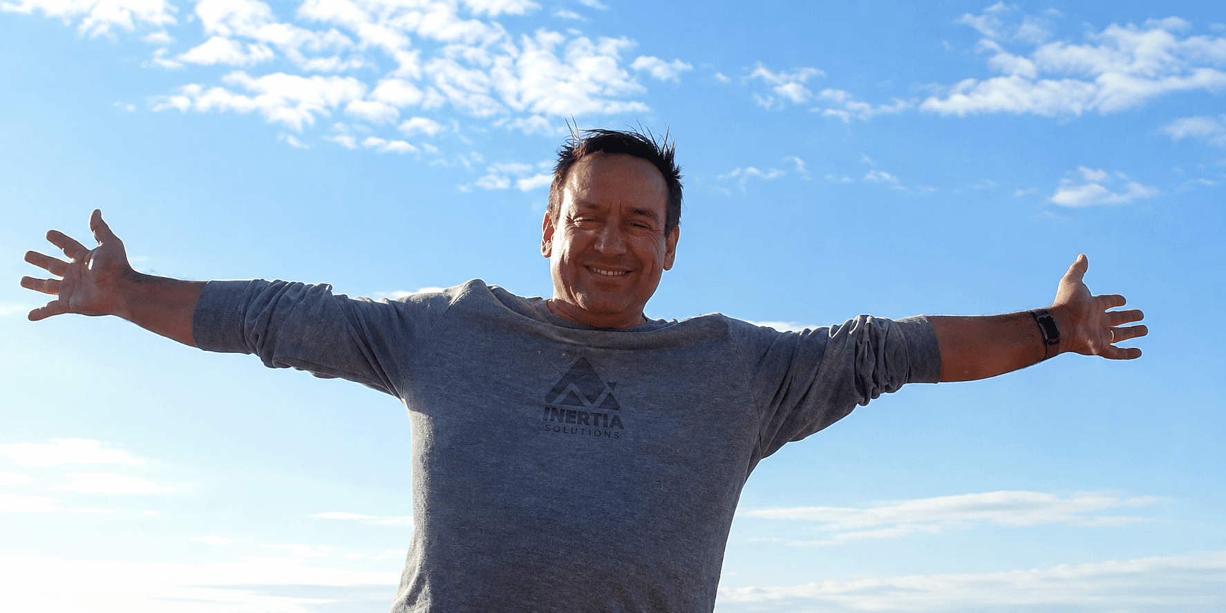 James Moss standing outdoors under an open sky, arms stretched wide, grinning, wearing an Inertia Solutions sweatshirt.